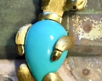 May include: A gold-toned poodle brooch with a turquoise blue cabochon stone. The poodle is standing on all four legs and has a green rhinestone eye.