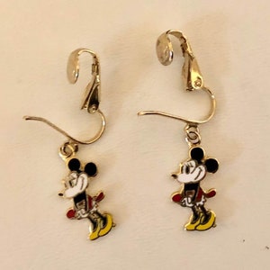 May include: A pair of gold-coloured clip-on earrings with a Mickey Mouse design. Each earring has a black head, white face, red jacket, and yellow shoes. The earrings are set against a light background.