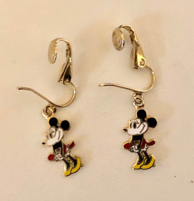 May include: A pair of gold-coloured clip-on earrings with a Mickey Mouse design. Each earring has a black head, white face, red jacket, and yellow shoes. The earrings are set against a light background.