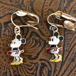 May include: A pair of gold-coloured clip-on earrings with a colourful Mickey Mouse design. The earrings have a black head, white face, red shorts, and yellow shoes. The clip-on mechanism is visible.