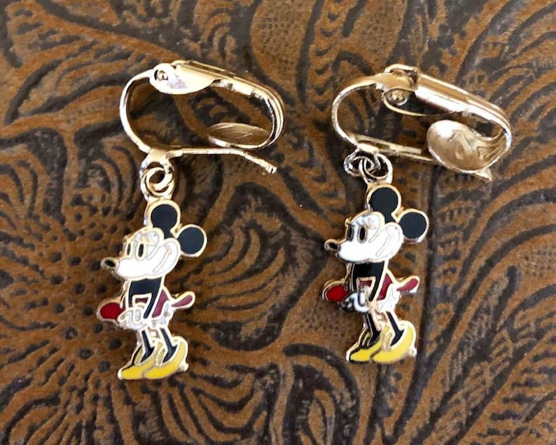 May include: A pair of gold-coloured clip-on earrings with a colourful Mickey Mouse design. The earrings have a black head, white face, red shorts, and yellow shoes. The clip-on mechanism is visible.