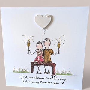 May include: A square greeting card featuring a line-art illustration of two figures sitting on a bench, holding gold glitter champagne flutes. Above them is a white heart balloon. The text reads: A lot can change in 30 years... but not my love for you.