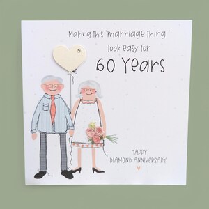May include: A square greeting card featuring an illustration of an older couple. The text reads: Making this 'marriage thing' look easy for 60 Years. HAPPY DIAMOND ANNIVERSARY. The couple holds a cream heart balloon and a small bouquet of pink flowers.