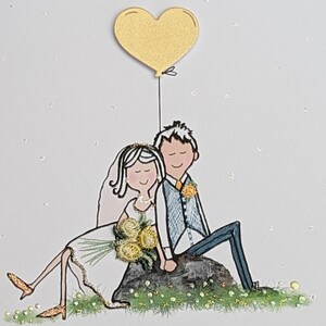 Może przedstawiać: Ilustracja na kartce ślubnej przedstawia parę młodą siedzącą na skale, trzymającą się za ręce. Panna młoda ma na sobie białą suknię i welon, trzyma bukiet. Złoty balon w kształcie serca unosi się nad nimi. Tło białe z brokatem.