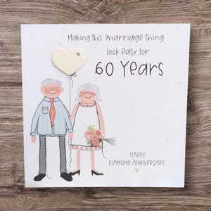 May include: A square greeting card featuring a cartoon illustration of an elderly couple. The text reads: Making this 'marriage thing' look easy for 60 Years. HAPPY DIAMOND ANNIVERSARY. The design includes a heart-shaped balloon and fabric flowers.