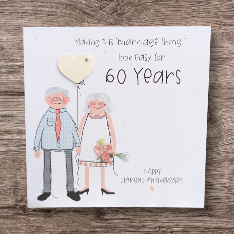 May include: A square greeting card featuring a cartoon illustration of an elderly couple. The text reads: Making this 'marriage thing' look easy for 60 Years. HAPPY DIAMOND ANNIVERSARY. The design includes a heart-shaped balloon and fabric flowers.