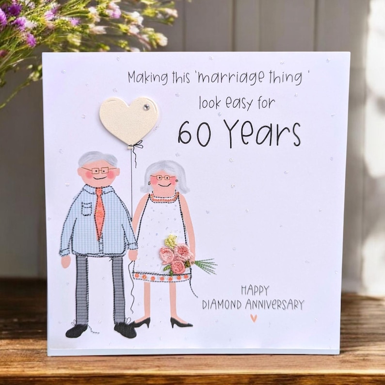 May include: A white greeting card featuring a cartoon illustration of an older couple. The text reads: Making this 'marriage thing' look easy for 60 Years. HAPPY DIAMOND ANNIVERSARY. The card includes a cream heart balloon and a small fabric flower bouquet.