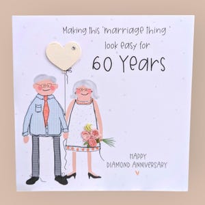 May include: A white greeting card featuring an illustration of an older couple with a heart-shaped balloon. The text reads: Making this 'marriage thing' look easy for 60 Years. HAPPY DIAMOND ANNIVERSARY. The couple wears embroidered clothing details.
