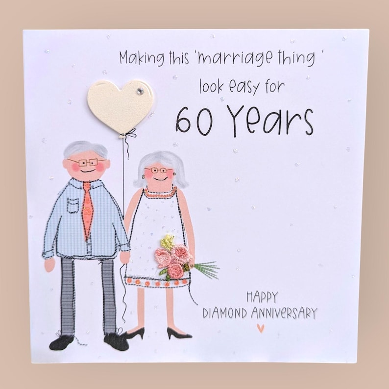 May include: A white greeting card featuring an illustration of an older couple with a heart-shaped balloon. The text reads: Making this 'marriage thing' look easy for 60 Years. HAPPY DIAMOND ANNIVERSARY. The couple wears embroidered clothing details.