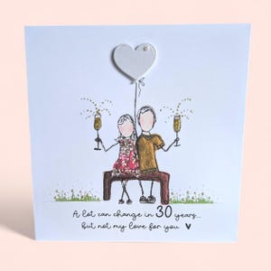 May include: A white greeting card featuring a hand-drawn illustration of two figures sitting on a brown bench, holding gold glitter champagne glasses. Above them is a white heart balloon. Text reads: A lot can change in 30 years... but not my love for you.