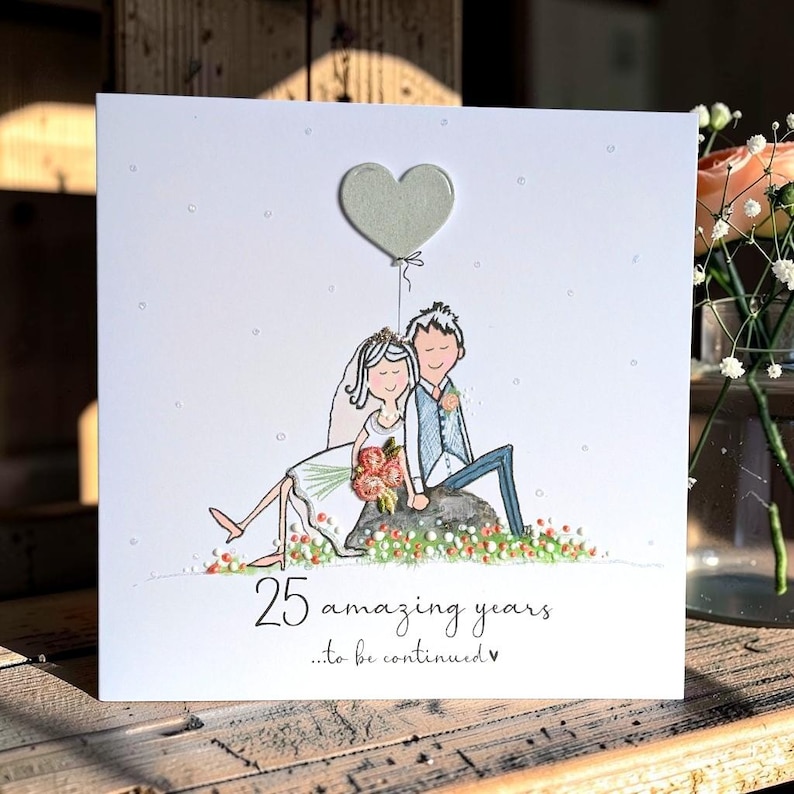 May include: A square white greeting card featuring a hand-drawn illustration of a couple sitting on a rock. The bride wears a white dress and veil, and the groom wears a blue vest and trousers. Above them is a heart-shaped balloon. Text reads 25 amazing years...to be continued.