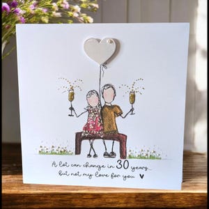 May include: A square greeting card featuring a hand-drawn illustration of two figures sitting on a bench, holding gold glitter champagne glasses. Above them is a white heart balloon. The text reads: A lot can change in 30 years... but not my love for you.