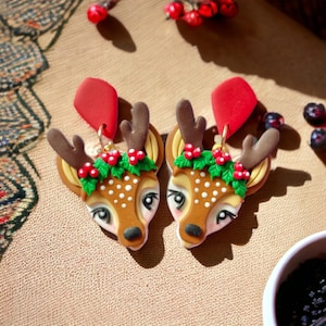 May include: A pair of deer head earrings. Each earring features a brown deer head with white spots, brown antlers, and a red accent. The deer heads are adorned with green holly and red berries, and are suspended from red diamond-shaped dangles.