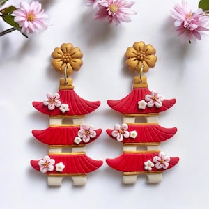 May include: A pair of red and gold pagoda earrings with white cherry blossom decorations. The earrings are made from polymer clay and have a gold flower charm at the top.