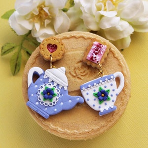 May include: A pair of handmade polymer clay earrings featuring a blue teapot and a blue teacup with white polka dots and a blue flower design. The earrings are attached to a biscuit-like charm with a red heart and a pink and white candy-like charm.