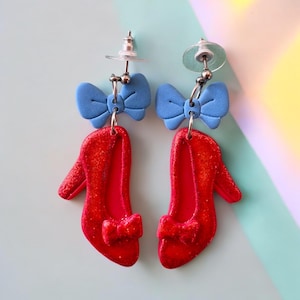 Könnte beinhalten: Handgefertigte Ohrringe mit roten Glitzer-High Heels und blauen Schleifen. Die Ohrringe haben silberfarbene Metallstifte. Die High Heels haben eine Schleife. Der Hintergrund ist pastellfarben.