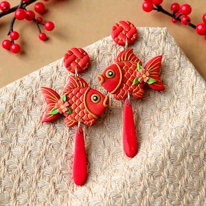 May include: A pair of red fish-shaped earrings with gold detailing and floral accents. Each earring features a round top and a long, teardrop-shaped dangle. The earrings are presented on a textured, beige surface.