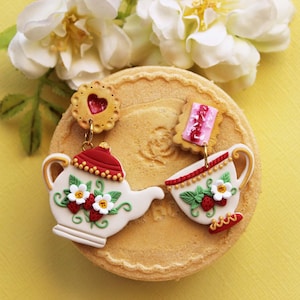 Könnte beinhalten: Ein Paar skurriler Ohrringe mit einem Teekannen- und Teetassen-Design, verziert mit Erdbeeren und Blumen. Die Ohrringe sind weiß mit roten und goldenen Details, vor einem gelben Hintergrund mit einem Keks-Sockel. Weiße Blumen sind im Hintergrund.