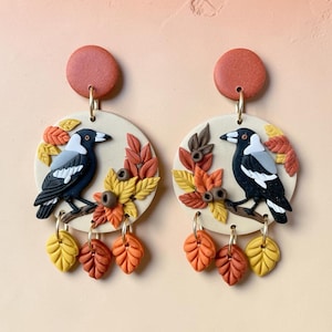 May include: A pair of drop earrings featuring a round terracotta-coloured stud connected to a larger cream-coloured circular disc. Each disc displays a black and white magpie perched on a branch surrounded by sculpted autumn leaves in shades of yellow, orange, and brown, with three additional leaf charms hanging from the bottom.