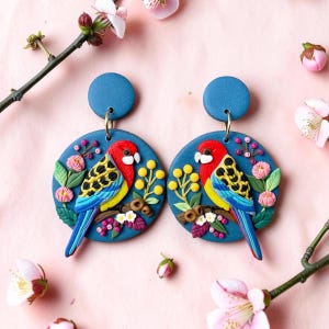 May include: A pair of circular blue dangle earrings featuring a detailed relief design of a colourful parrot with red, yellow, and blue feathers, perched on a branch surrounded by small pink flowers, green leaves, and yellow berries.