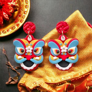 May include: A pair of red, blue and yellow lion dance earrings with gold accents. The earrings are hanging from a gold fabric background.