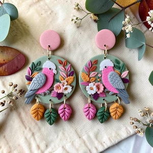 May include: A pair of dangle earrings featuring circular sage green discs with sculpted pink and grey galah cockatoos, colourful floral accents, and three small leaf-shaped charms in yellow, green, and pink hanging from the bottom edge.