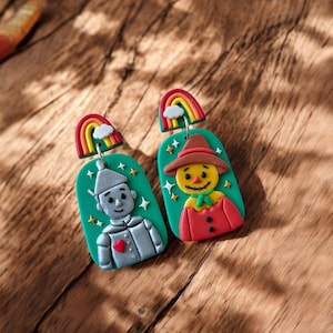 May include: A pair of novelty earrings featuring characters from The Wizard of Oz. One earring shows the Tin Man with a red heart, the other the Scarecrow. Each earring has a green background with stars and a rainbow with a cloud.