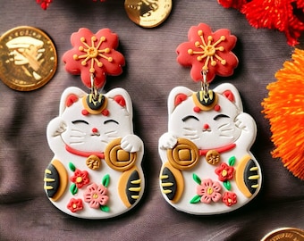 May include: A pair of white lucky cat earrings with red and pink flowers, gold coins, and black accents. The earrings are made of polymer clay.