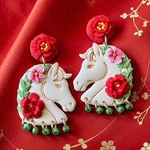 May include: A pair of white horse head earrings with red and pink floral details and green bead dangles. The earrings have gold-coloured hardware, set against a red background.