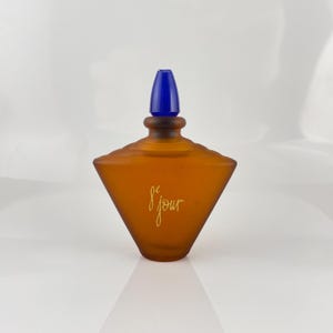 以下が含まれることがあります： 幾何学的なデザインと青いストッパーが付いた琥珀色の香水瓶。 ボトルには、前面に金色のスクリプトで「de jour」という文字が書かれています。 ボトルは白い表面にあります。