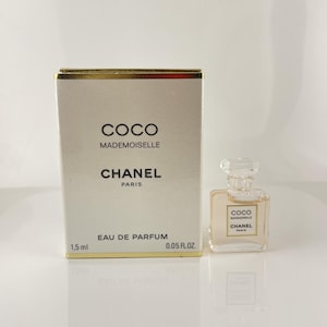 Pode incluir: Um frasco em miniatura do perfume Coco Mademoiselle Chanel ao lado da sua caixa a condizer. A caixa e o frasco são bege claro com detalhes dourados. A caixa diz "COCO MADEMOISELLE CHANEL PARIS" e "EAU DE PARFUM 1,5 ml 0,05 FL.OZ."