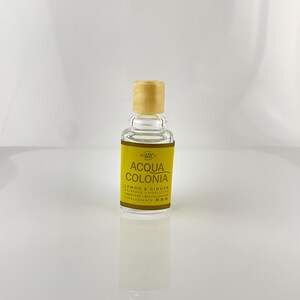 Pode incluir: Uma pequena garrafa de vidro transparente de Acqua Colonia Limão e Gengibre. A garrafa tem um rótulo amarelo com texto preto e uma tampa de madeira castanha clara. O rótulo inclui a marca e detalhes do produto.