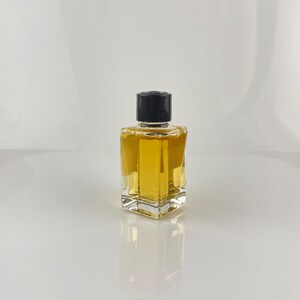 May include: A small, square perfume bottle with a black cap. The bottle is filled with a golden-colored liquid. The glass bottle has a geometric design and is sitting on a reflective surface.