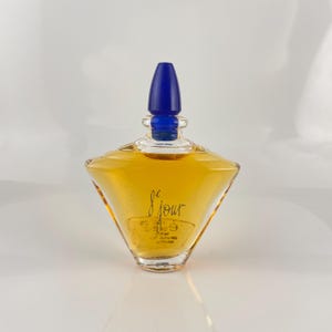 Puede incluir: Un frasco de perfume de vidrio transparente con un tapón azul. El frasco tiene una forma geométrica y contiene un líquido de color dorado. Las palabras "Je Jour" están impresas en la parte delantera del frasco.
