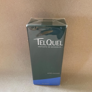 Puede incluir: Una caja rectangular de aftershave, envuelta en plástico transparente. La caja es de color verde oscuro con la palabra "TELQUEL" en blanco, y también se ve el texto "INSTANTS DE BONHEUR..." y "APRES-RASAGE". La parte inferior de la caja es azul.