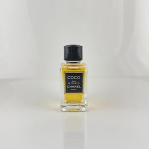 May include: A small, rectangular perfume bottle with a black cap. The bottle contains a golden-yellow liquid and features a black and gold label with the text "COCO EAU DE TOILETTE CHANEL PARIS."