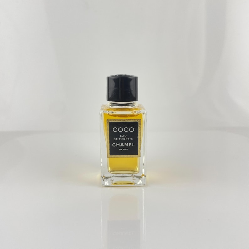May include: A small, rectangular perfume bottle with a black cap. The bottle contains a golden-yellow liquid and features a black and gold label with the text "COCO EAU DE TOILETTE CHANEL PARIS."