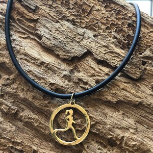 Puede incluir: Un collar dorado con un colgante que presenta una silueta recortada de un corredor. El collar está en un cordón negro y está descansando sobre una pieza de madera.