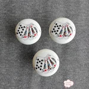 May include: Three white ceramic knobs with a design of playing cards on the top. The cards are arranged in a fan shape and include the Jack, Queen, King, and Ace of Spades, as well as the Queen of Hearts.