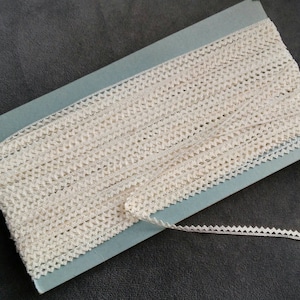 Puede incluir: Un rollo de encaje de ganchillo de color blanco roto con un borde festoneado. El encaje está en una tarjeta azul.