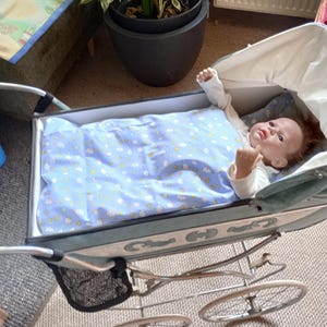 Op de afbeelding: Een vintage kinderwagen in lichtblauw en zilver, met spaakwielen en een zwart opbergmandje. Een pop ligt erin, bedekt met een blauwe deken met witte wolkenpatronen. De kinderwagen heeft een witte kap.