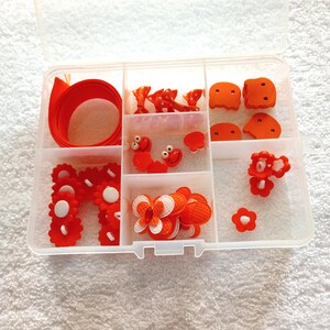 May include: A clear plastic storage box with multiple compartments filled with orange and white craft supplies. The box contains ribbon, bows, flowers, buttons, and other embellishments.