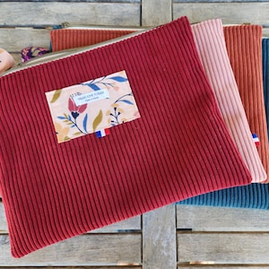 Può includere: Una collezione di pochette con cerniera in velluto a coste nei toni del rosso, rosa, arancione e verde acqua. Le pochette hanno una texture a coste e una toppa floreale rettangolare con testo. Un nastro fantasia è attaccato alla cerniera.