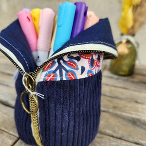 Peut inclure: Une trousse en velours côtelé bleu marine avec une fermeture éclair dorée, contenant des marqueurs colorés. L'intérieur présente un tissu à motifs. Les marqueurs sont roses, jaunes, rose clair, bleus, violets et pêche.