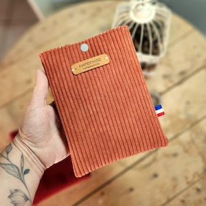 Peut inclure: Une pochette rectangulaire en velours côtelé couleur rouille avec des rayures verticales. Elle est dotée d'un bouton-pression argenté au centre supérieur et d'une étiquette en similicuir beige cousue avec le texte HANDMADE limited edition.