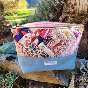 Può includere: Un beauty case in tessuto trapuntato con un design patchwork multicolore. La borsa ha una base in denim azzurro e una fodera rosa a quadretti. La cerniera è dorata e presenta un'etichetta con la scritta "HINAE AIME FLOWER".