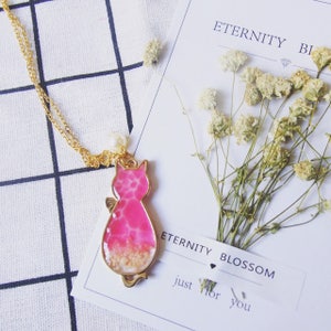 Puede incluir: Un collar de cadena dorada con un colgante de gato rosa relleno de purpurina y flores secas. El colgante tiene un acento de perla. El collar se muestra sobre un fondo blanco con una tarjeta que dice "Eternity Blossom just for you".