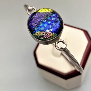May include: A silver bangle bracelet with a round, iridescent glass cabochon. The glass is blue, purple, and yellow with a swirling pattern.