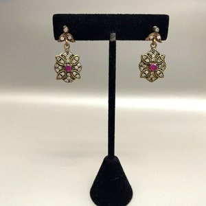 May include: A pair of gold-toned earrings with a floral design. Each earring features a central red stone surrounded by small, clear stones. The earrings are displayed on a black stand against a white background.