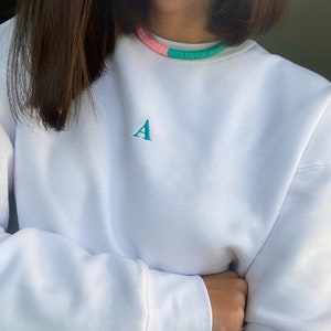 Può includere: Felpa bianca con una piccola lettera 'A' ricamata sul davanti. La felpa ha una collana di perline rosa e turchese.